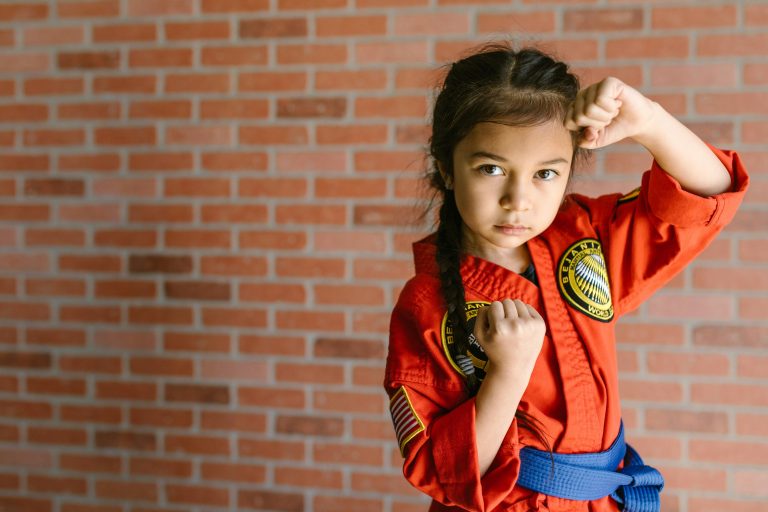 A determined young girl in a karate uniform practices martial arts indoors against a brick wall.