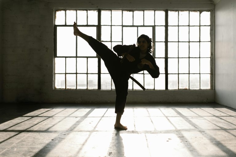 Martial artist performing a high kick in a sunlit room creating dramatic shadows.